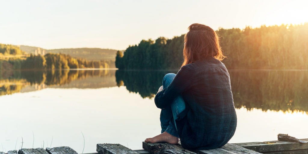 Kvinna sedd bakifrån sitter på brygga vid en blank sjö, tema självmedkänsla.