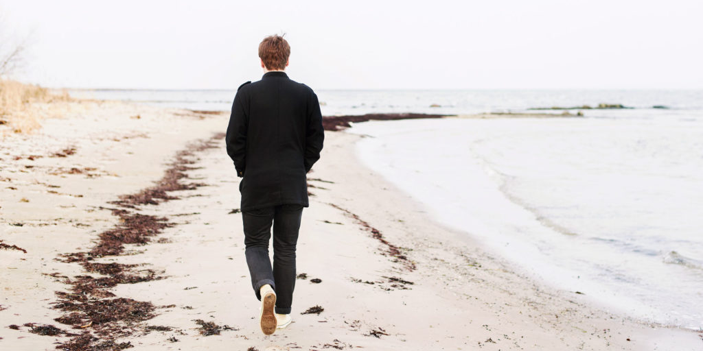 En man i jacka och långbyxor går på en höstlik strand