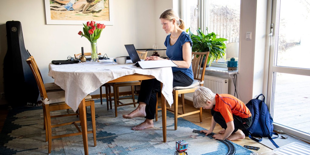 Kvinna som arbetar framför dator vid matsalsbord hemma. BArm leker med tågbana under bordet.