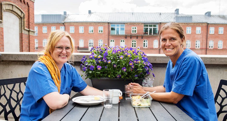 Två personer i blå vårdpersonalkläder sitter och äter matlådelunch utomhus.