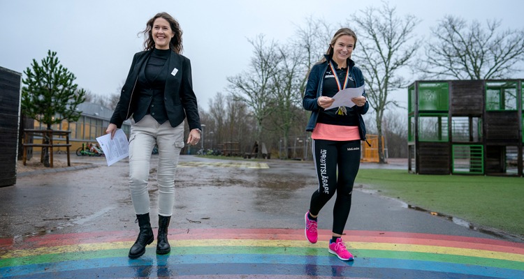Två kvinnor går bredvid varandra över en skolgård. På marken har man målat en regnbåge. I bakgrunden syns en klätterställning.