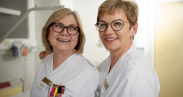 Två kvinnor i vårdpersonalkläder tittar mot kameran och ler