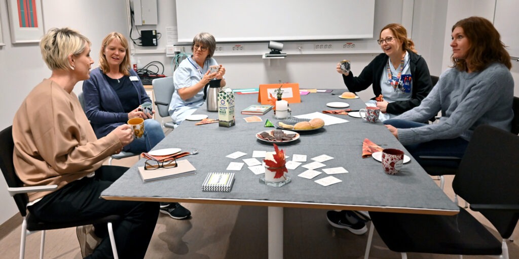 Fem kvinnor sitter runt ett bord och fikar och samtalar