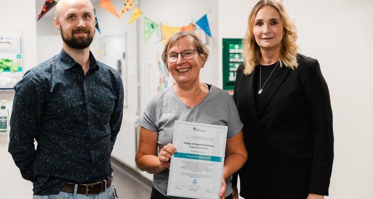 Jonatan Dahlbo, Petra Ludvigson och Raija Hållinggård på Holmens dagliga verksamhet står i en korridor. Petra Ludvigson håller i en tavla med utmärkelsen årets hygienombud.