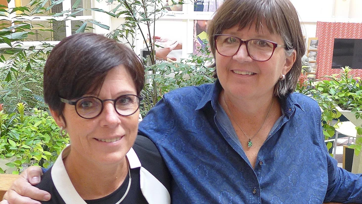 susanne hylander, upphandlare, och agneta kallstenius, arbetsmiljöcontroller, ler mot kameran och står tätt ihop i en ljushall med grönska bakom ryggen.