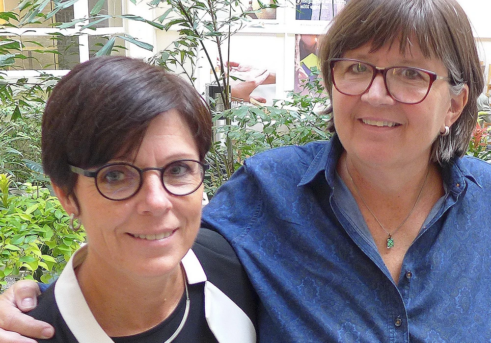 susanne hylander, upphandlare, och agneta kallstenius, arbetsmiljöcontroller, ler mot kameran och står tätt ihop i en ljushall med grönska bakom ryggen.