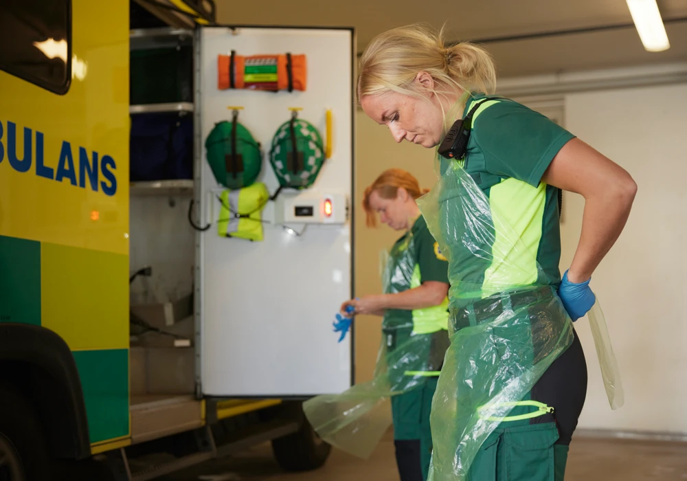 Två kvinnliga ambulanssjukvårdare tar på plastförkläden utanför en ambulans, tema ia-systemet för incidentrapportering.