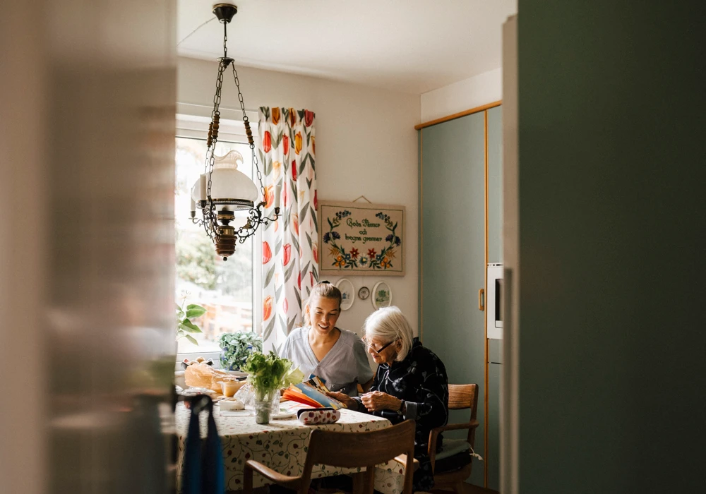 Personal från hemtjänsten och en äldre person sitter i ett kök och pysslar med något tillsammans.