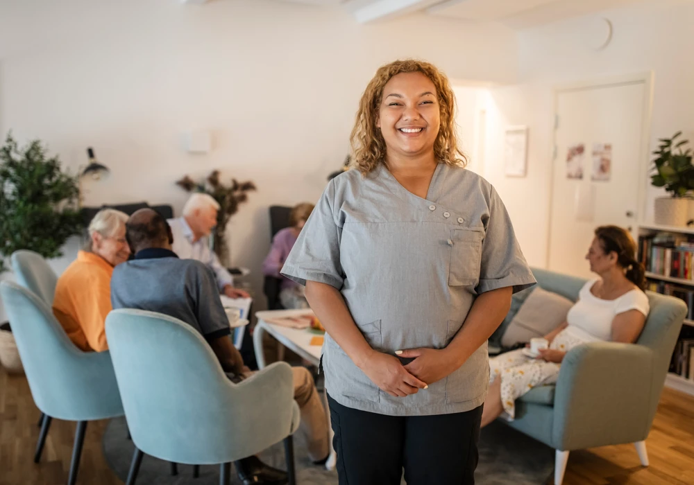 En leende kvinna i arbetskläder för omsorg, timanställd inom äldreomsorgen, står framför en grupp människor som sitter i soffor och pratar med varandra.