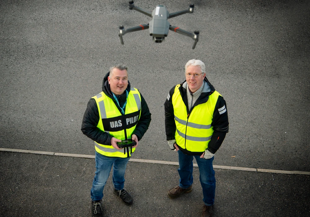 Två män i varselvästar står bredvid varandra framför en flygande drönare.
