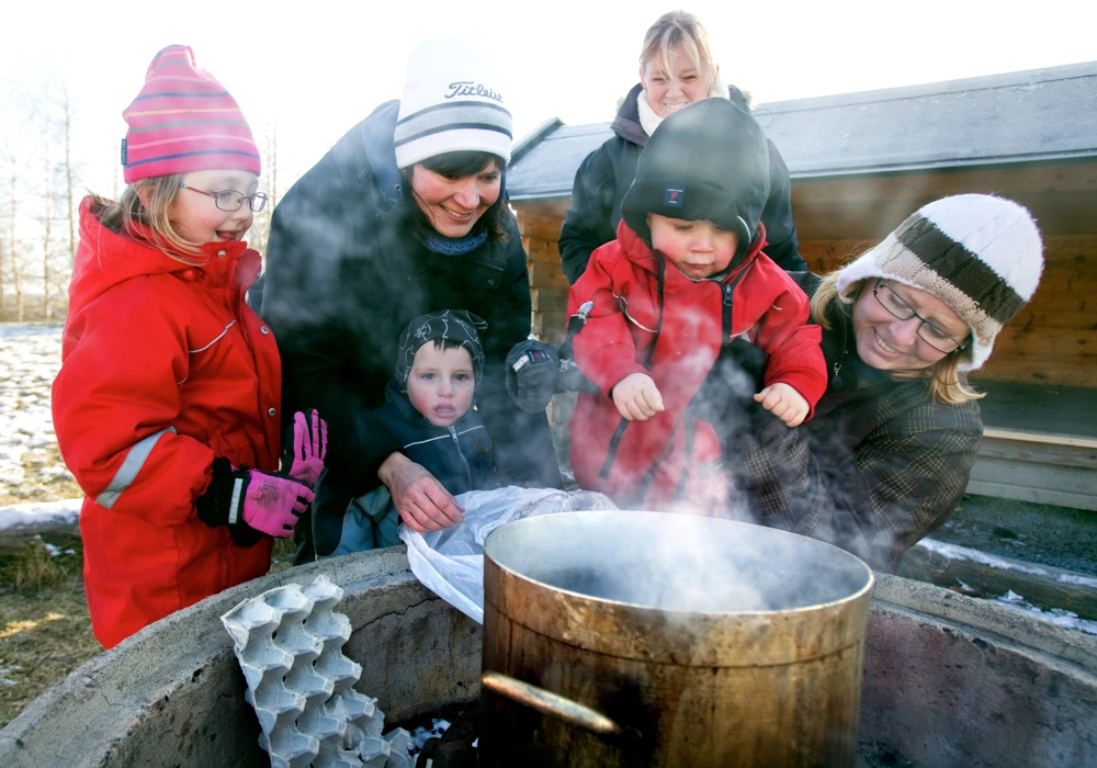 En grupp små barn och vuxna kvinnor som står utomhus runt en kastrull med kokande vatten.