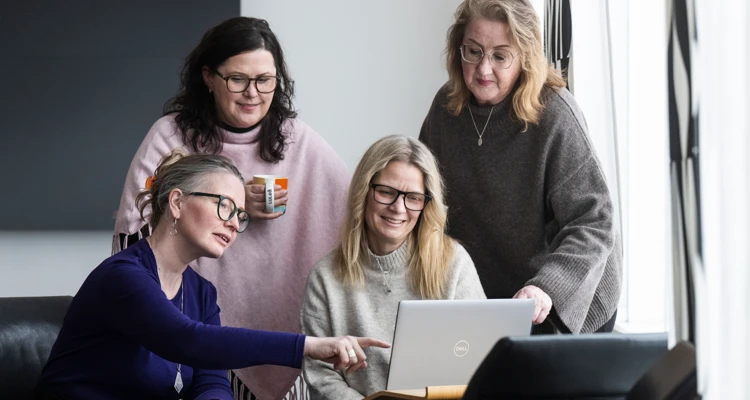En grupp kvinnor som sitter och står runt en bärbar dator med vackert ljus från ett fönster.