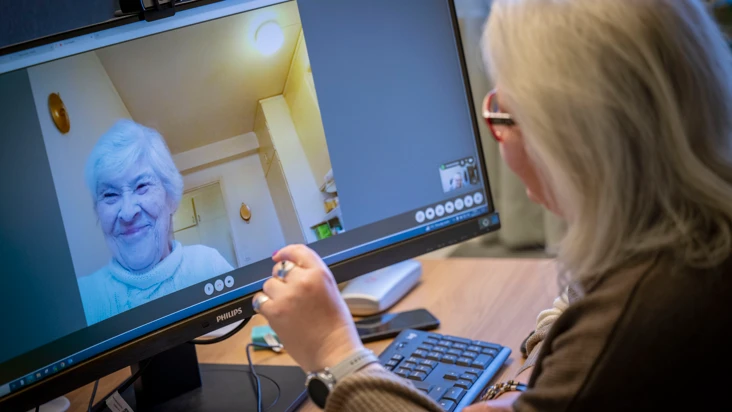 En kvinna från hemtjänsten sitter framför en datorskärm och pratar med en brukare.