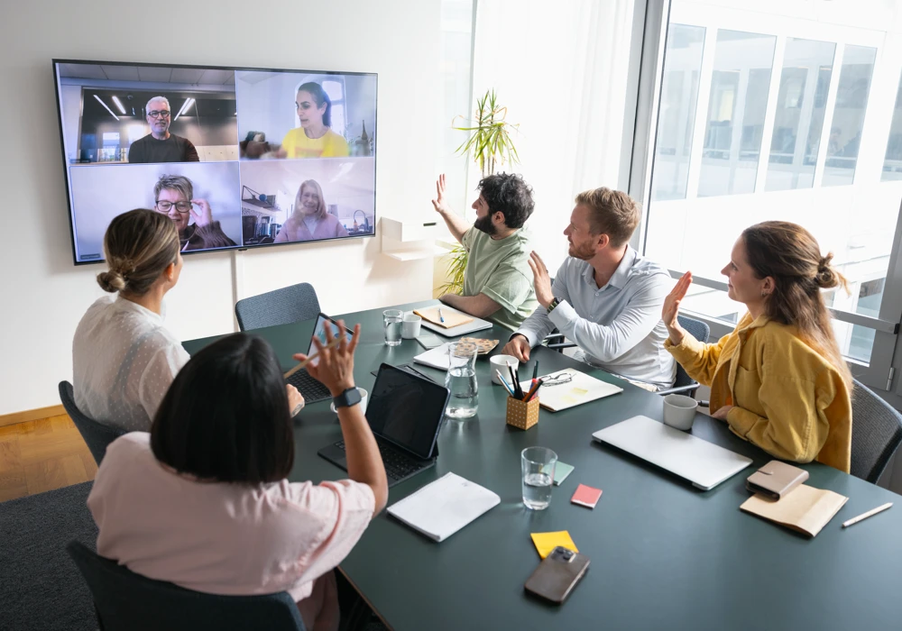 En grupp människor som sitter runt ett konferensbord och har ett hybridmöte med människor på en skärm.