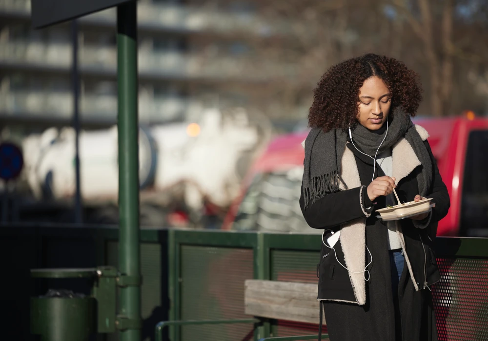 En kvinna som står vid en busshållplats och äter matlåda, med mobilens hörlurar i öronen.