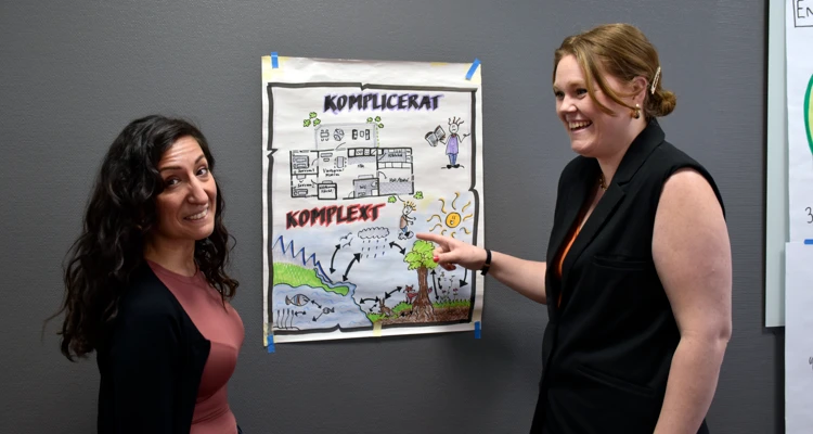 Två kvinnor står vid en vägg där det hänger ett blädderblocksblad med illustrationer.