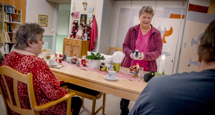 Hemtjänstmedarbetare står vid bord och håller dekoration, vid bordet sitter några personer.