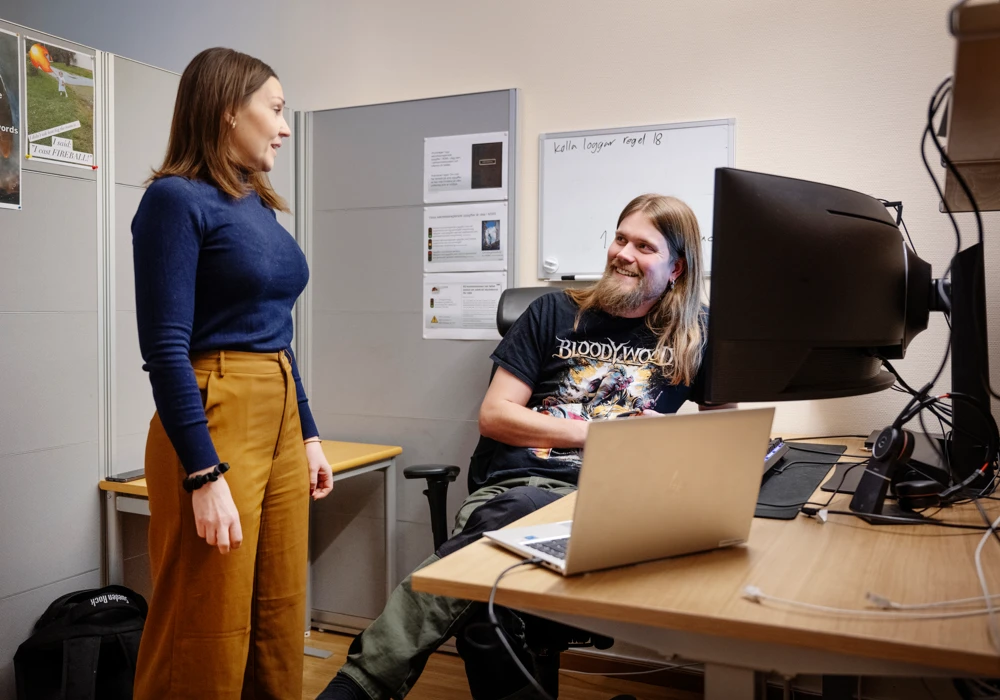 Kontorsrum med en man som sitter vid skrivbord med datorer, kvinna som står.