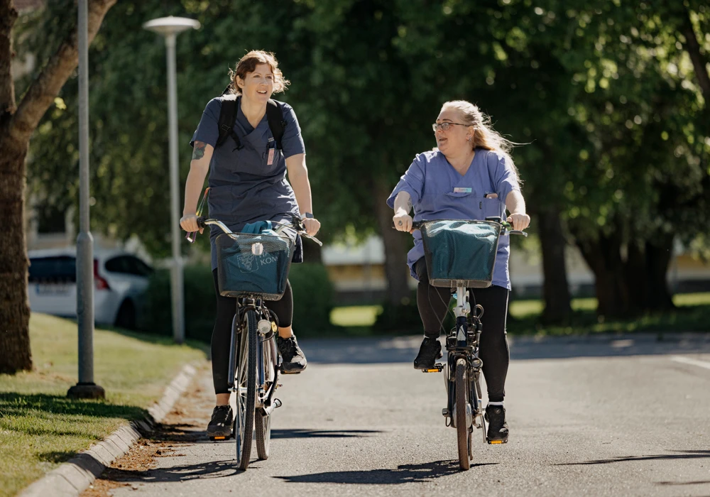 två kvinnliga undersköterskor i blå arbetskläder cyklar på mindre väg med grönska i bakgrunden.