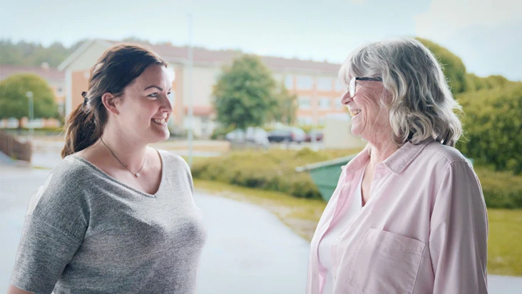 Två leende kvinnor som står utomhus och pratar med varandra.